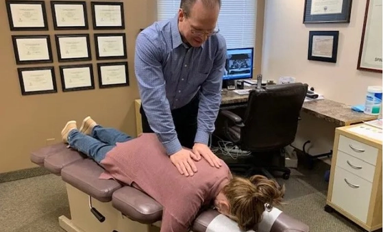 A person is lying face down on a padded chiropractic table while another person applies pressure to the back. The room has framed certificates on the wall, a desk with a computer, and office furniture