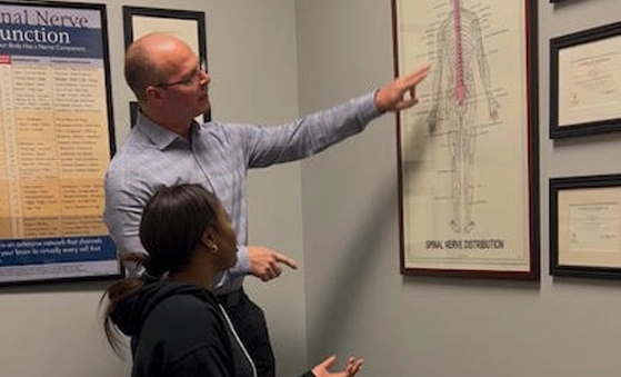 Dr. Aaron Williams pointing at a framed anatomical chart of spinal nerve distribution on a wall, with another person observing