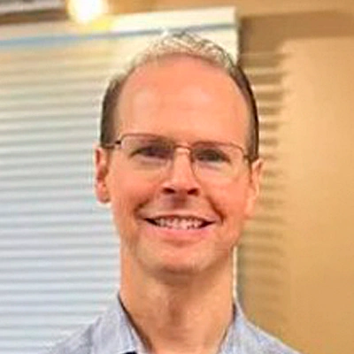 Dr. Aaron Williams wearing a light blue collared shirt standing indoors with a background featuring horizontal blinds and a beige wall