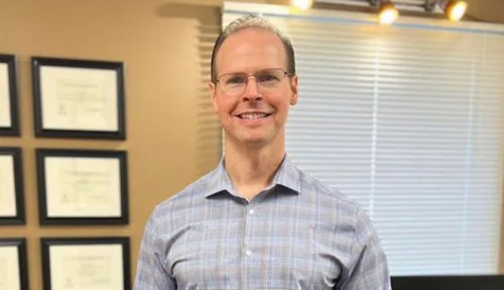 Dr. Aaron Williams wearing a light gray checkered button-up shirt standing indoors, with framed certificates on the wall and a window with blinds in the background